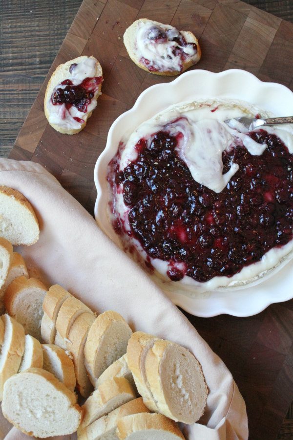 Baked Brie with Blueberry Sauce recipe from RecipeGirl.com #brie #blueberry #summer #appetizer #recipe #RecipeGirl overhead shot of melted baked brie in a white dish with basket of baguette slices on the side and baguette slices with some of the brie and blueberry sauce on them- all sitting on a wooden cutting board