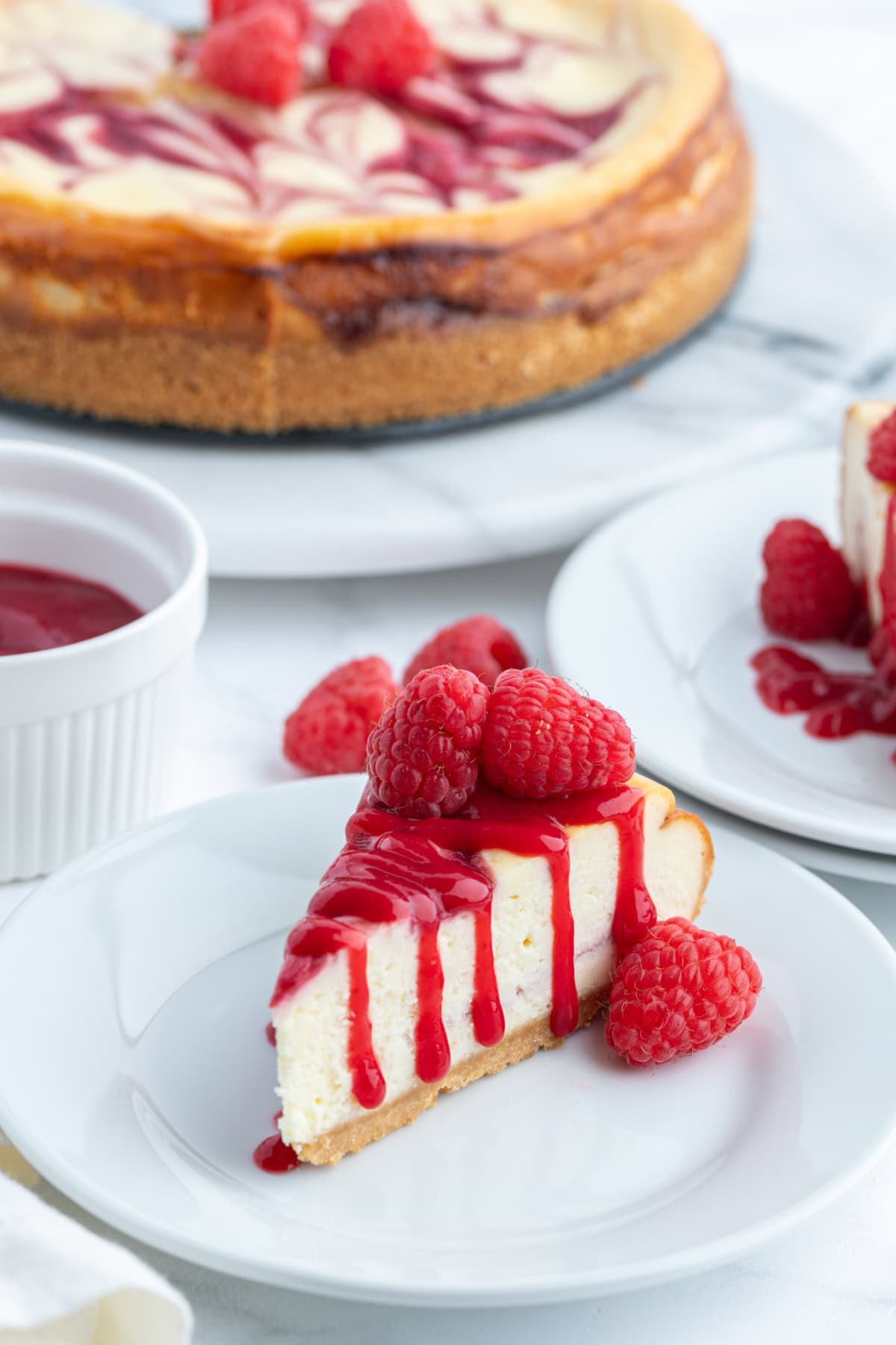 White Chocolate Raspberry Cheesecake recipe from RecipeGirl.com slice of white chocolate raspberry cheesecake on plate with whole cheesecake in background
